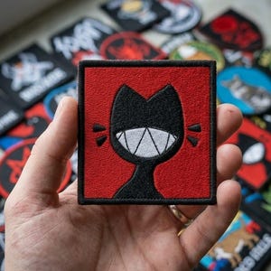 May include: A square embroidered patch featuring a black cat silhouette with a wide, toothy grin on a vibrant red background. The patch has a thick black border and is held up against a background of various other assorted embroidered patches.