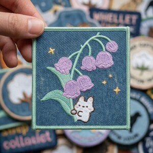 May include: A square denim patch featuring an embroidered design of lily of the valley flowers in shades of purple and green, with a small white bunny and gold stars. The patch has a green border and is held in a hand, with other patches in the background.
