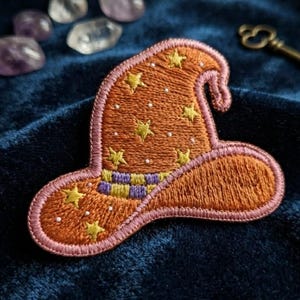 May include: An embroidered patch shaped like a pointed hat in burnt orange with a pink border. The hat features yellow embroidered stars, small white dots, and a purple and yellow checkered band around the base, all stitched with visible thread.