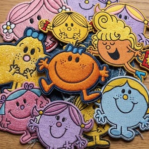 May include: A collection of embroidered fabric patches featuring various colorful cartoon characters with distinct shapes, hair styles, and facial expressions, including round, star-shaped, and oval figures in shades of pink, yellow, orange, purple, and blue.