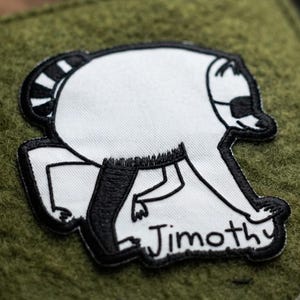 May include: An olive green felt bag featuring a white embroidered patch of a stylized raccoon character. The raccoon has black outlines, a striped tail, and a black mask. The name Jimothy is embroidered in black script below the character.