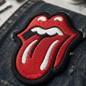 May include: An embroidered patch featuring a red, white, and black graphic of lips with a tongue sticking out. The patch is stitched onto a dark blue denim fabric, showcasing detailed thread work with a thick black border outlining the design.
