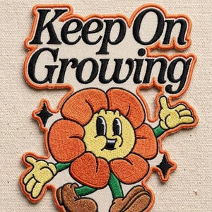 May include: An embroidered patch featuring the text Keep On Growing in black letters with an orange border. Below the text is a cartoon flower character with orange petals, a yellow face, green arms, and brown shoes, accented by two black stars.