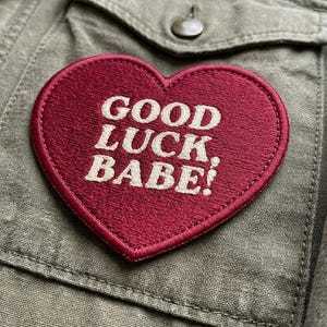 May include: A heart-shaped embroidered patch in deep red with the words "GOOD LUCK BABE!" in white lettering. The patch is affixed to a green-gray jacket, with a pocket and button visible in the background.