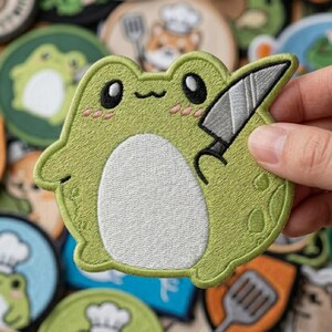 May include: Embroidered patch featuring a cartoon frog holding a knife. The frog is light green with a white belly and pink cheeks. The patch is surrounded by other embroidered patches with various designs.