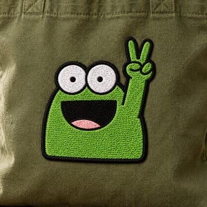 May include: An olive green canvas tote bag featuring a large, embroidered patch of a bright green cartoon frog character with wide white eyes, a black mouth, and a raised arm making a peace sign gesture, centered on the front of the bag.