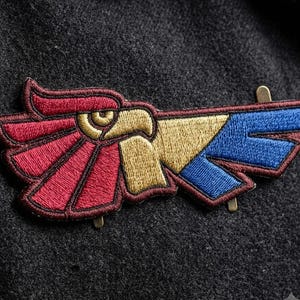 May include: An embroidered patch featuring a stylized eagle head profile in gold, with wings depicted in segmented sections of deep red and blue. The design is outlined in dark thread and includes two small metal attachment pins on the back side.