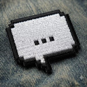 May include: A rectangular embroidered patch featuring a white pixelated speech bubble design with a black border and three black square dots in the center, representing a typing indicator icon, attached to a textured blue denim fabric surface.