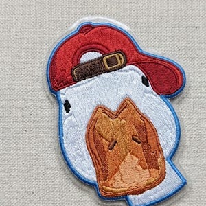 May include: Embroidered patch featuring a cartoon duck wearing a red baseball cap with a brown buckle. The duck has a white head, a brown beak, and is outlined in blue. The patch is designed for application on clothing or accessories.