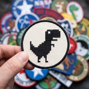 May include: A round, off-white patch featuring a black pixelated dinosaur design. The patch is held in front of a blurred background of various other patches with different designs and colors. The dinosaur is facing left.