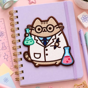 May include: A light purple spiral-bound notebook featuring an embroidered patch of a tan cat dressed as a scientist. The cat wears round glasses and a white lab coat, holding a green liquid flask in one paw and a pink liquid flask in the other.