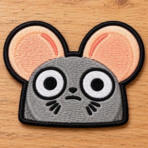 May include: An embroidered patch featuring a gray mouse head with large, round white eyes, black pupils, and a small black nose. The mouse has prominent, rounded peach-colored ears with a black border and three black whiskers on each cheek.