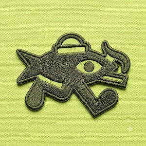 May include: An embroidered patch featuring a dark olive green stylized fish shape with a prominent central eye, a small rectangular top handle, and a smoking cigarette extending from the right side, all outlined in a thick matching thread border.