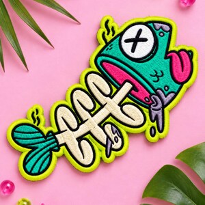 May include: An embroidered patch featuring a cartoon fish skeleton with a teal head, a white eye with a black X, a pink tongue, and purple accents. The fish has a cream-colored bone structure and a teal tail, all outlined in bright neon yellow thread.