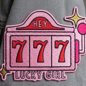 May include: A pink embroidered patch in the shape of a slot machine with the number 7 in red three times. The patch has the words "HEY" and "LUCKY GIRL" in red and pink. The patch is on a gray denim jacket.