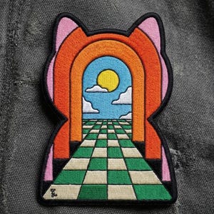 May include: An embroidered patch shaped like a cat head featuring an orange archway leading to a blue sky with a yellow sun and white clouds. The foreground shows a green and beige checkered floor. The design is outlined in black thread.