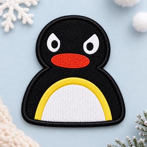 May include: An embroidered patch shaped like a penguin character with a black body, a white belly outlined in yellow, a bright orange beak, and white eyes with black pupils. The patch features a dense, textured stitch pattern throughout.