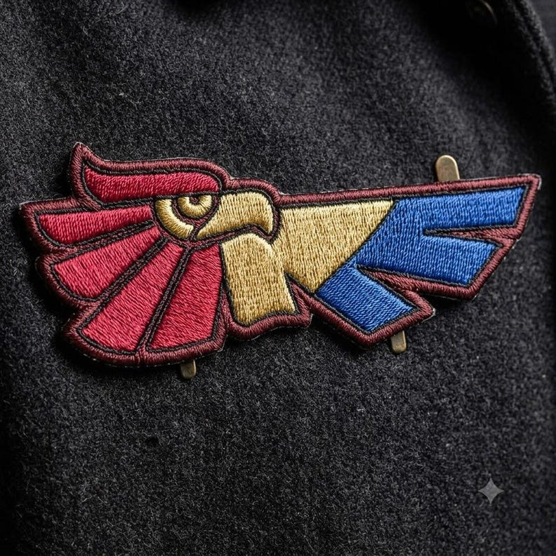 May include: An embroidered patch featuring a stylised eagle head profile in gold, with wings depicted in segmented sections of deep red and blue. The design is outlined in dark thread and includes two small metal attachment pins on the back side.