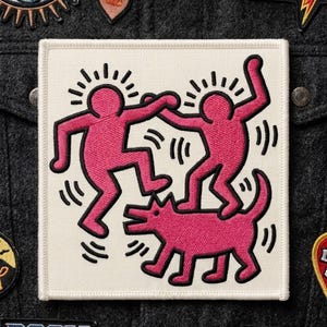 May include: A square patch featuring a pink Keith Haring-style design of two figures dancing above a dog. The figures and dog are outlined in black on a white background. The patch is surrounded by other patches on a dark gray fabric.