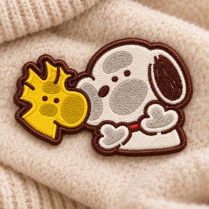 May include: An embroidered patch featuring a white dog with gray spots and a red collar, positioned next to a small yellow bird character. The patch has a dark brown stitched border and is placed on a textured cream-colored knit fabric.