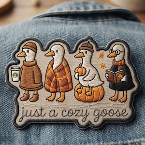 May include: An embroidered patch featuring four geese in autumn attire, including one with a coffee cup, one in a plaid blanket, one on a pumpkin, and one reading a book. The text "just a cozy goose" is stitched at the bottom in dark thread.