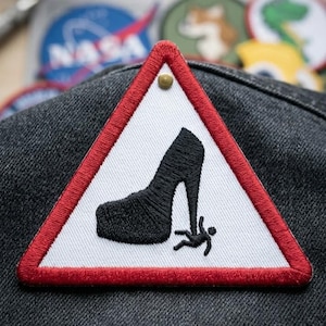 May include: Embroidered patch featuring a black high-heeled shoe stepping on a stick figure, set within a red-bordered white triangle. A small gold-colored tack secures the patch. The patch is on a dark gray denim-like fabric.