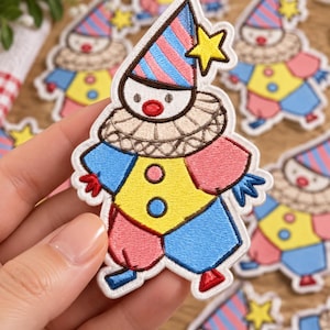 May include: An embroidered clown patch featuring a pointed hat with blue and pink stripes, a yellow star, a ruffled collar, and a colorful suit in yellow, blue, and pink. The clown has a white face with a red nose and two small blue buttons.