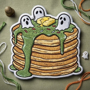 May include: An embroidered patch featuring a stack of golden brown pancakes topped with green syrup, colorful sprinkles, a yellow pat of butter, and three small white ghost figures with black eyes and mouths, all outlined in white thread.