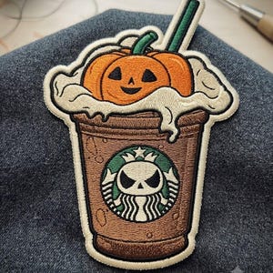 May include: An embroidered patch featuring a brown coffee cup with a green circular logo containing a white skull character. The cup is topped with white whipped cream, a small orange jack-o'-lantern pumpkin, and a green straw sticking out.
