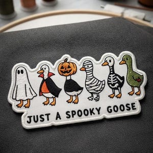 May include: An embroidered patch on dark gray fabric featuring six geese in Halloween costumes: a ghost, a vampire, a pumpkin-headed goose, a mummy, a skeleton, and a green goose. The text "JUST A SPOOKY GOOSE" is embroidered in black below.