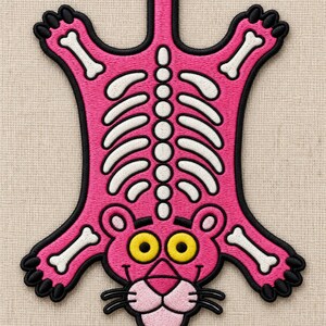May include: An embroidered patch featuring a stylized pink panther figure with a visible white skeletal structure. The design includes yellow eyes, a pink snout, black whiskers, and a black border outline on a textured light beige fabric background.