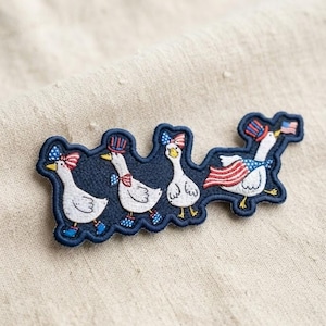 May include: An embroidered patch featuring four white geese on a dark blue background. The geese are decorated with red, white, and blue patriotic motifs, including star-spangled hats, bows, boots, and a cape, with one goose holding a small American flag.