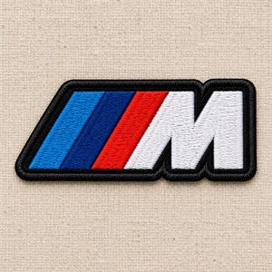May include: An embroidered patch featuring the letter M in white, preceded by three slanted stripes in light blue, dark blue, and red. The entire design is outlined with a thick black border, creating a distinct shape with a flat top and bottom.