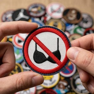 May include: A round patch with a white background, a black bra crossed out with a red line, and a red border. The patch is held between two hands, with other patches in the background.