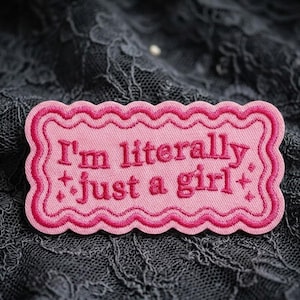 May include: A pink embroidered patch with the phrase "I'm literally just a girl" in pink lettering. The patch has a wavy border and star accents. The background is a dark gray lace fabric.
