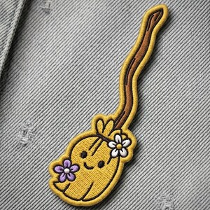 May include: An embroidered patch shaped like a yellow broomstick with a smiling face. The broom has a long brown handle, a yellow bristle base, a white flower on the right side, and a purple flower on the left side, all outlined in yellow thread.