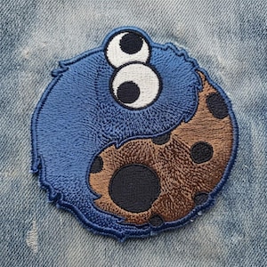 May include: A circular embroidered patch featuring a blue fuzzy character with two large white eyes and black pupils, arranged in a yin-yang style with a brown chocolate chip cookie shape that has dark brown circular chips throughout the design.