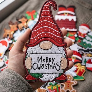 May include: An embroidered patch featuring a gnome with a long white beard, a tall red hat with white polka dots, and red boots. The gnome holds a white rectangular sign with the black text Merry Christmas printed in a handwritten style font.
