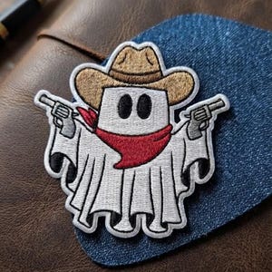 May include: An embroidered patch featuring a white ghost character wearing a tan cowboy hat and a red bandana around its neck. The ghost has two black oval eyes and is holding a grey revolver in each of its two outstretched arms.