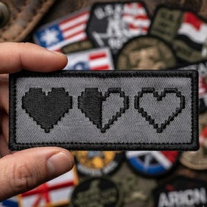 May include: A rectangular gray patch with a black embroidered border featuring three pixelated heart icons. The first heart is solid black, the second heart is half black and half outlined, and the third heart is a black outline.