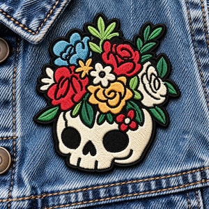 May include: An embroidered patch featuring a cream-colored skull with black eye sockets and a nose cavity. A vibrant bouquet of red, yellow, white, and blue flowers with green leaves emerges from the top of the skull, all outlined in black thread.