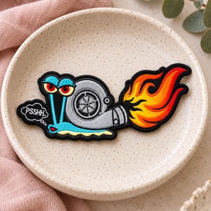 May include: An embroidered patch featuring a light blue snail with red eyes and a grey turbocharger shell. The snail has a speech bubble containing the text PSSHH and a trail of orange and yellow flames extending from the back of the turbocharger.
