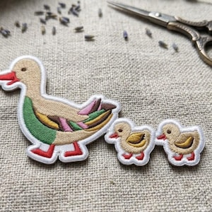 May include: An embroidered patch featuring a large duck with a tan head, green chest, and multi-colored wing feathers, followed by two smaller ducklings. Each figure has a white border, a tan body, a red beak, and red feet.