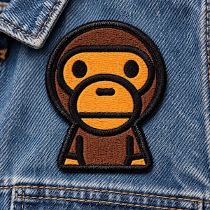 May include: An embroidered patch featuring a stylized brown and yellow monkey character with a black outline, attached to a blue denim jacket. The monkey has a round face, large black eyes, and a simple expression, rendered in dense thread work.