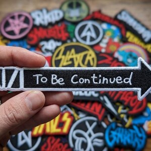 May include: Black and white embroidered patch in the shape of an arrow, with the text "To Be Continued". The patch is held in a hand, with other patches in the background.