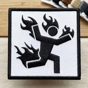 May include: A square embroidered patch featuring a white background with a thick black border. The design depicts a black stick figure in a running pose, with flames emanating from the head, hands, and one leg, all rendered in black thread.