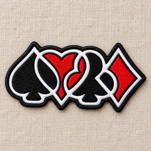 May include: An embroidered patch featuring the four playing card suits arranged in a row. The design includes a black spade, a red heart, a black club, and a red diamond, all outlined in thick white and black thread borders on a fabric base.