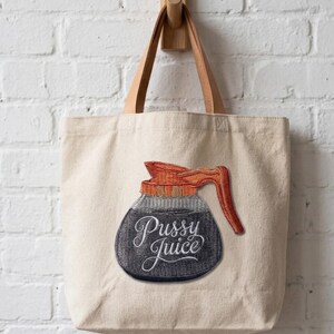 May include: A beige canvas tote bag with brown leather handles hanging on a wooden peg. The bag features an embroidered patch of a coffee pot with the words "Pussy Juice" in cursive. The coffee pot is black, gray, and orange.