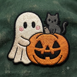 May include: An embroidered patch featuring a white ghost with pink cheeks, a small gray cat, and a bright orange jack-o'-lantern with a black carved face. The design is outlined in black thread and set against a dark green fabric background.