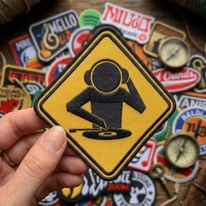 May include: A yellow and black embroidered patch featuring a DJ graphic within a diamond shape. The DJ is depicted with headphones and a turntable. Other patches and a compass are in the background.